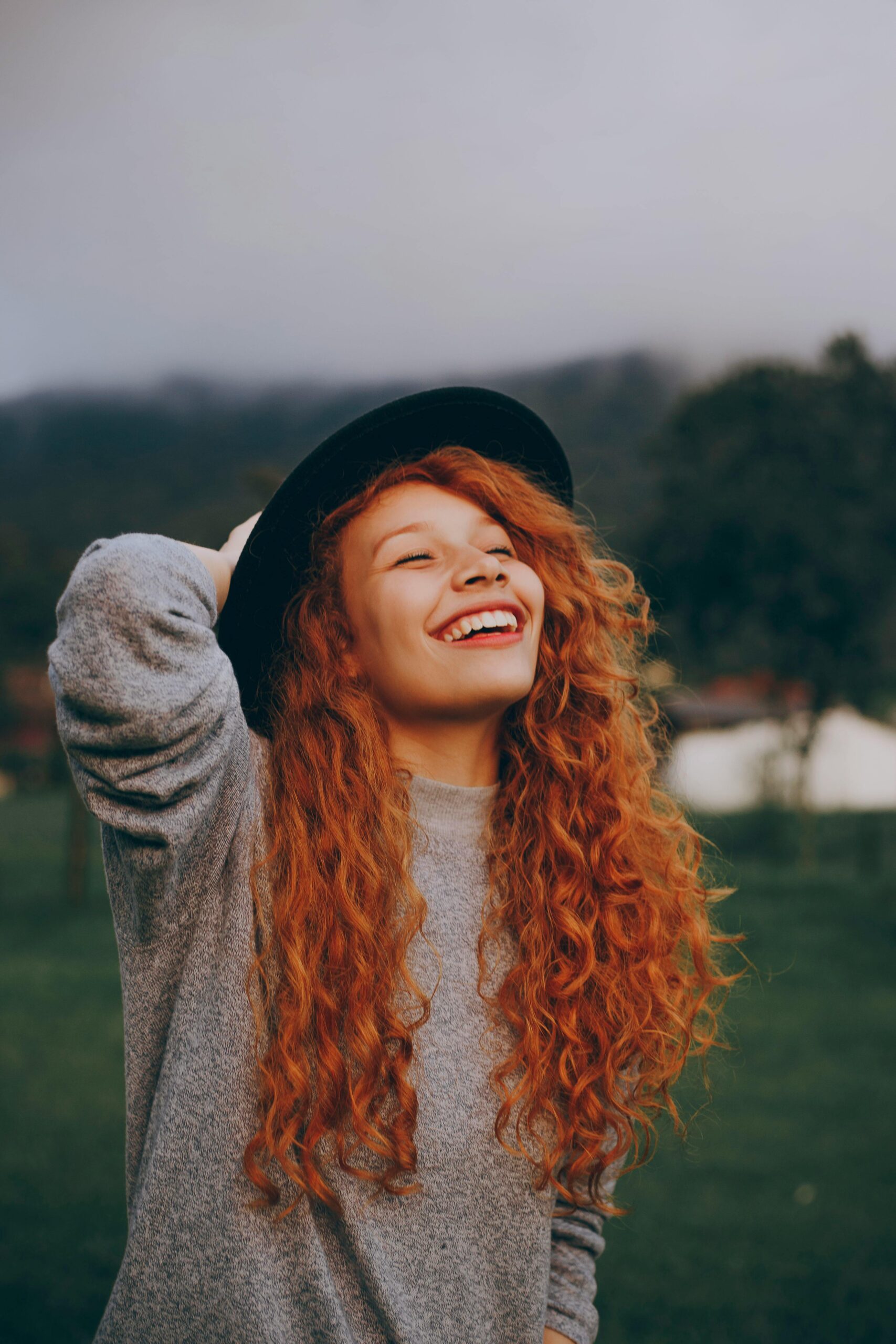 A happy young woman with curly red hair and a hat smiling in a lush green park.
