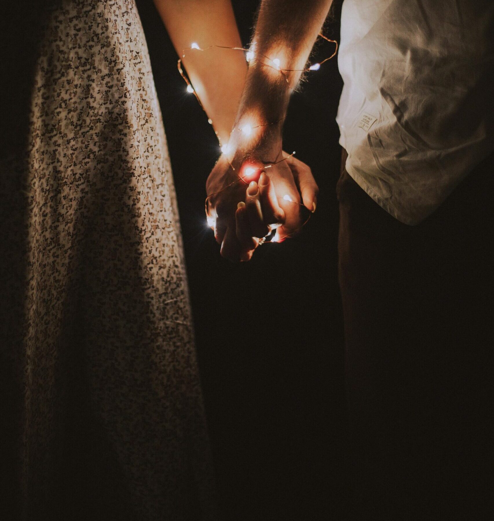 A warm and intimate close-up of a couple holding hands adorned with glowing string lights.